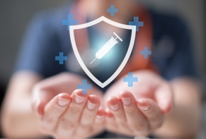 Manos abiertas en gesto de protección con un gráfico digital de un escudo con una jeringa en el centro, simbolizando la vacunación como método de prevención y cuidado de la salud.
